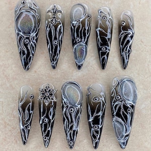 Op de afbeelding: Een set van tien lange, puntige kunstnagels. Elke nagel heeft een zwart en zilver ontwerp met ingewikkelde patronen en versieringen. De nagels hebben een glanzende afwerking en zijn gerangschikt op een lichtgekleurd oppervlak. De tekst "Nail Art" staat bovenaan.