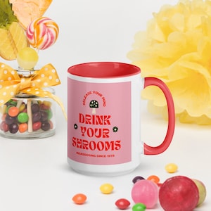 May include: A white ceramic mug with a red rim and handle. The mug has a pink background with the text "Release Your Mind Drink Your Shrooms Microdosing Since 1979" and a green and red mushroom illustration.