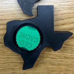 May include: Two black, Texas-shaped containers. One is a solid shape, the other has a circular opening with the text "GEO- CACHE" in green. The containers are on a wooden surface.