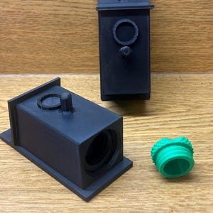 May include: Black 3D-printed birdhouse components, including a rectangular body with a circular opening, a separate roof piece, and a green, textured plug. The birdhouse parts are displayed on a wooden surface.