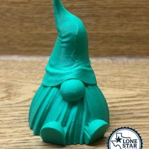 May include: A teal-colored gnome figurine with a pointed hat and round belly. The gnome is sitting with its legs extended. The figurine is made of a solid material and has a textured surface. The Lone Star Cache Supply logo is in the bottom right corner.