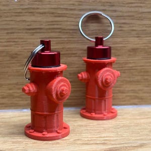May include: Two red fire hydrant keychains with silver key rings. The keychains have a detailed design, including the top cap, side nozzles, and base. The keychains are made of a 3D-printed material and sit on a wooden surface.