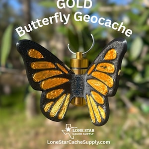 May include: A gold and black butterfly geocache with the text "GOLD Butterfly Geocache". The butterfly has gold-colored accents on its wings and a gold-colored container. The geocache is from Lone Star Cache Supply.