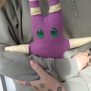 May include: A person is holding a purple and yellow plush toy with green eyes. The toy has a star shape and is made of soft fabric. The person has several paw print tattoos on their arm.