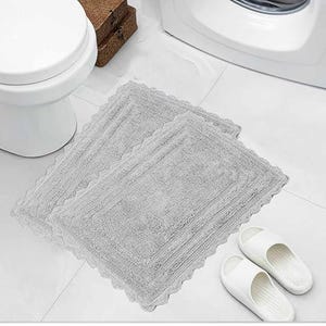 May include: Two light gray rectangular bath mats with a scalloped edge are placed on a white tiled floor. White sandals are next to the mats. A white toilet and a brown box are also in the image.