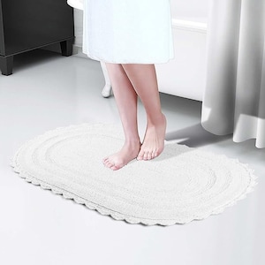 May include: White oval bath mat with a scalloped edge. The mat is made of a soft, absorbent material and is placed on a light-colored floor. The mat is designed to be placed in front of a bathtub.