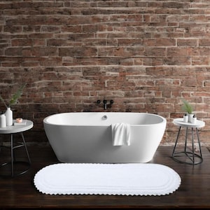 Könnte beinhalten: Eine weiße, ovale Badematte mit gewelltem Rand liegt auf einem dunklen Holzboden vor einer weißen Badewanne. Das Badezimmer hat eine Backsteinmauer und zwei kleine Beistelltische.