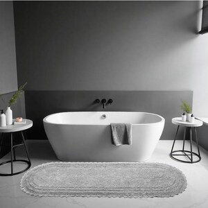 Könnte beinhalten: Ein modernes Badezimmer mit einer weißen ovalen Badewanne, einer grauen Badematte und zwei kleinen Beistelltischen. Die Wände sind in Grautönen gestrichen, was eine minimalistische und ruhige Atmosphäre schafft. Ein graues Handtuch hängt am Wannenrand.