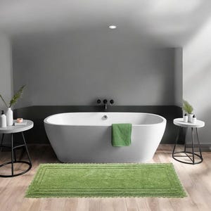 Könnte beinhalten: Ein modernes Badezimmer mit einer weißen ovalen Badewanne, einer grünen Badematte und zwei kleinen Beistelltischen. Ein grünes Handtuch hängt an der Wanne. Die Wände sind grau und der Boden ist helles Holz.