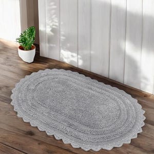 May include: A grey, oval bath mat with a scalloped edge. The mat is made of a soft, absorbent material and is placed on a wooden floor. A small potted plant is visible in the background.