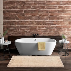 Könnte beinhalten: Eine beige Badematte mit dekorativem Rand vor einer weißen Badewanne in einem Badezimmer mit einer Backsteinwand.