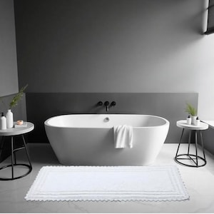 Könnte beinhalten: Ein modernes Badezimmer mit einer weißen ovalen Badewanne, einer weißen Badematte mit gewelltem Rand und zwei kleinen Beistelltischen. Die Wände sind in Grautönen gestrichen. Ein weißes Handtuch hängt am Wannenrand.