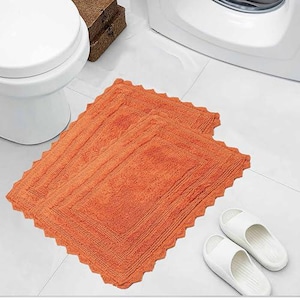 May include: Two orange rectangular bath mats with a scalloped edge are displayed on a white tiled floor. White sandals are next to the mats. A white toilet and a small brown box are in the background.