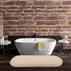 Könnte beinhalten: Ein modernes Badezimmer mit einer weißen ovalen Badewanne, einem gelben Handtuch, einer beigen ovalen Badematte und zwei kleinen Beistelltischen. Der Hintergrund ist eine Backsteinmauer, die eine warme Atmosphäre schafft.