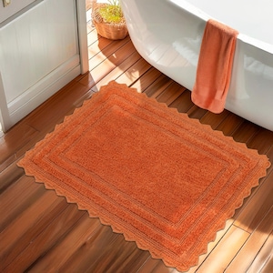 Puede incluir: Una alfombra de baño rectangular naranja con borde festoneado sobre un suelo de madera. La alfombra tiene una superficie texturizada y un borde más oscuro. Una toalla naranja cuelga del borde de una bañera blanca.
