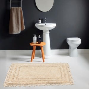 Könnte beinhalten: Ein beiger Badezimmerteppich mit einer gehäkelten Bordüre. Der Teppich liegt vor einem weißen Waschbecken auf einem Sockel und einer weißen Toilette.