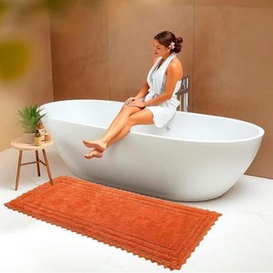 Könnte beinhalten: Eine ovale weiße Badewanne mit einer Person, die am Rand sitzt und einen weißen Bademantel trägt. Eine rostfarbene rechteckige Badematte mit gewelltem Rand liegt auf dem Boden. Ein kleiner Beistelltisch mit einer Pflanze und Flaschen befindet sich in der Nähe.