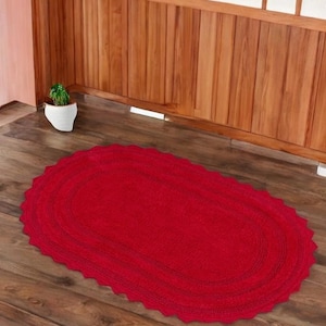 May include: A vibrant red, oval rug with a decorative, scalloped edge. The rug is made of a textured material and is placed on a wooden floor. A small potted plant is in the background.