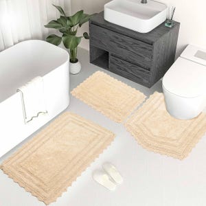 Puede incluir: Un juego de tres alfombras de baño beige con borde de ganchillo. Las alfombras se colocan delante de una bañera blanca, un inodoro y un tocador con un lavabo.