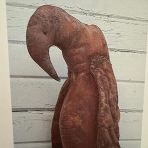 May include: A brown, textured, soft sculpture shaped like a curved, elongated figure with a prominent, downward-pointing hook at the top. The surface has a mottled, uneven appearance with visible stitching along the sides of the vertical body.