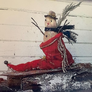 May include: A fabric snowman figure wearing a small hat and a dark scarf, nestled inside a long red stocking. The snowman has twig arms and a sprig of greenery attached. The stocking is positioned on a wooden base with a curled toe and a bell.