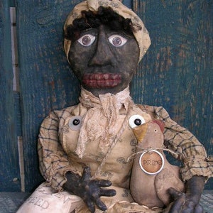 May include: A handmade doll with a dark complexion, wearing a patterned dress and head covering. The doll holds a small, wooden chicken with a "PRIDE" tag and a bag that says "FEED". The doll's features include large eyes and a wide mouth.