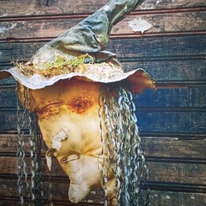 May include: A fabric doll head with a pointed hat, featuring long, thin, gray yarn hair, a small nose, and a safety pin detail. The hat is dark green with a frayed brim and moss-like accents. The doll has a light beige, textured surface.