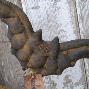 May include: A handcrafted, distressed-look bat decoration in shades of brown and black. The bat has a textured surface and is made of a soft material. A small tag is attached with twine. Perfect for Halloween or gothic decor.