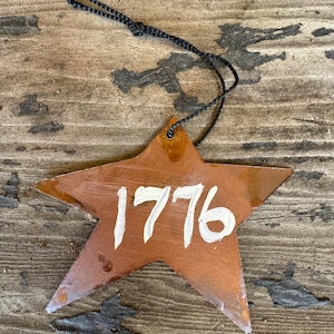 May include: A rusty orange star-shaped ornament with a black string for hanging. The number 1776 is painted in white on the star.