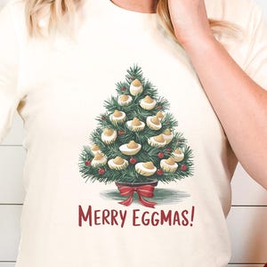 Peut inclure: T-shirt crème avec un motif d'arbre de Noël fait d'œufs mimosa et un nœud rouge. Le texte "Merry Eggmas!" est imprimé en dessous. Ce t-shirt festif fait partie d'une collection de vêtements de vacances.