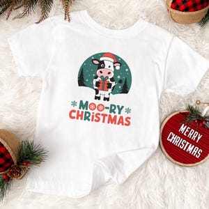 May include: White t-shirt with a black and white cow wearing a Santa hat and holding a gift. The text "*MOO-RY* CHRISTMAS" is printed on the shirt.