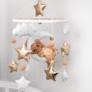 May include: A baby mobile featuring a lion cub resting on a crescent moon, surrounded by gold and white stars, clouds, and peach-colored spheres. The mobile is suspended from a white circular frame and a white mechanical device.