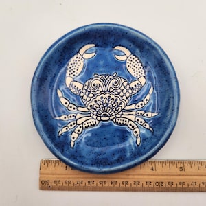 May include: A round, blue ceramic dish with a crab design. The crab is white with intricate patterns and is centered on the dish. The dish is approximately 10 cm in diameter.