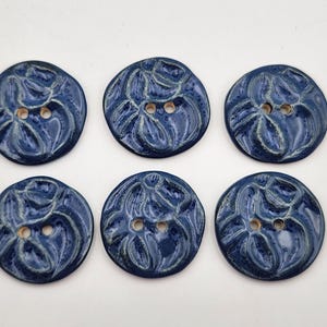 Puede incluir: Seis botones de cerámica hechos a mano en un esmalte azul oscuro con un diseño floral. Cada botón tiene dos agujeros para coser. Los botones son redondos y tienen una forma ligeramente irregular, lo que añade atractivo artesanal.