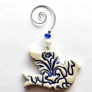May include: A white ceramic dove ornament with blue floral designs. The ornament has a silver wire hanger with a white pearl and blue beads.