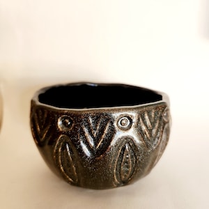 May include: A small, dark green ceramic bowl with a decorative pattern of leaves and circles around the rim.