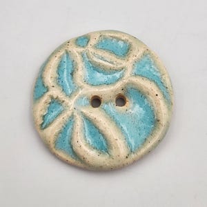 May include: A round ceramic button with a light blue floral design and two holes. The button has a cream-colored border and a speckled texture. The floral design is raised and has a glossy finish.