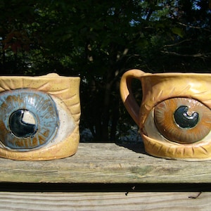 May include: Two ceramic mugs with eye designs. One mug has a blue eye with a brown iris and the other has a brown eye with a black iris.