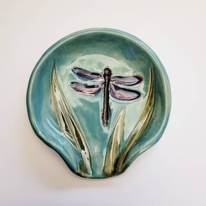 May include: A teal ceramic spoon rest with a dragonfly design. The spoon rest features a raised dragonfly with purple and black accents, and stylized green leaves. The spoon rest is a functional and decorative kitchen accessory.