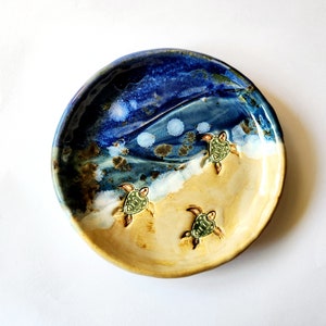 May include: A ceramic dish with a blue and yellow glaze, depicting a beach scene with three sea turtles. The dish is perfect for holding jewelry or other small items.