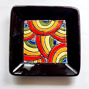 May include: A black ceramic tray with a colourful geometric design. The design features a pattern of overlapping semi-circles in shades of red, yellow, green, blue, and purple.