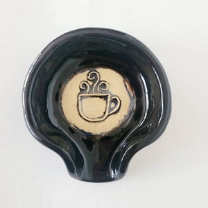 May include: Black ceramic spoon rest with a cream-coloured circle in the centre featuring a stylised black coffee cup with steam rising from it.