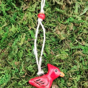 May include: A red ceramic cardinal bird pendant with a white string and a red bead. The bird has a small orange beak and is set against a background of green moss.