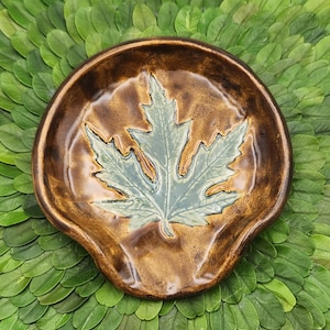 May include: A ceramic spoon rest with a maple leaf design. The spoon rest is brown with a light blue leaf in the center. The spoon rest is on a bed of green leaves.
