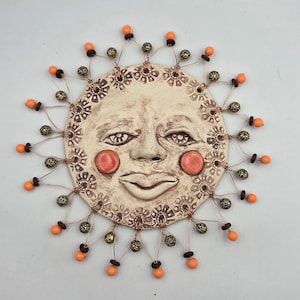 May include: A ceramic sun face wall hanging with a cream-colored base, featuring a smiling face with orange cheeks and a decorative border. The sun's rays are represented by orange beads and gold-colored beads attached with copper wire.