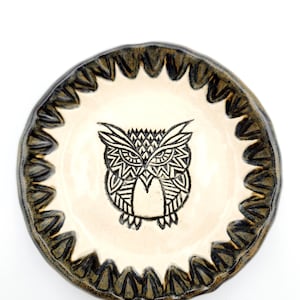 May include: A white ceramic dish with a black and white owl design. The dish has a scalloped edge and a dark green glaze.