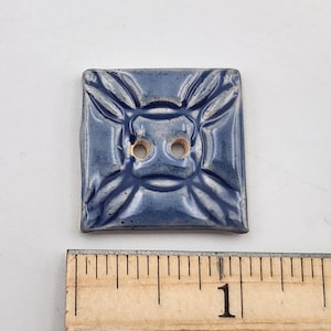 May include: Square ceramic button with a blue glaze and a raised floral design. The button has two holes for sewing and measures approximately 1 inch square.