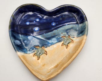 May include: Heart-shaped ceramic dish with a beach and ocean design. The dish features a gradient of colors, including dark blue, light blue, and tan. Two turtle designs are placed on the tan section. The dish is likely used for decorative purposes.