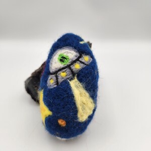 May include: A blue felt ball with an embroidered image of a spaceship and stars. The spaceship has a green window and yellow lights.