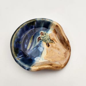May include: A blue and brown ceramic spoon rest with a sea turtle design. The spoon rest is handmade and has a unique, organic shape.